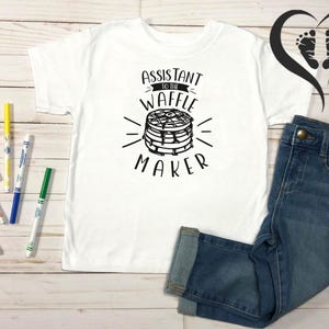 Puede incluir: Camiseta blanca con el texto "Assistant to the Waffle Maker" y un gr&aacute;fico de gofres. La camiseta se combina con vaqueros azules y rotuladores de colores. Un logotipo en forma de coraz&oacute;n est&aacute; en el fondo.