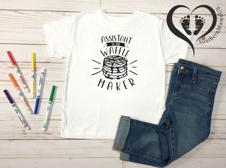 Puede incluir: Camiseta blanca con el texto "Assistant to the Waffle Maker" y un gr&aacute;fico de gofres. La camiseta se combina con vaqueros azules y rotuladores de colores. Un logotipo en forma de coraz&oacute;n est&aacute; en el fondo.