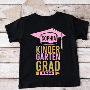 May include: Black t-shirt with a pink graduation cap design and the name "Sophia". The text "Kindergarten Grad" is in pink and gold. A pencil graphic is at the bottom.