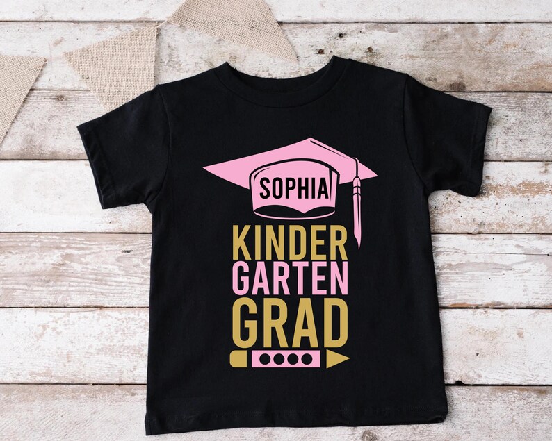 May include: Black t-shirt with a pink graduation cap design and the name "Sophia". The text "Kindergarten Grad" is in pink and gold. A pencil graphic is at the bottom.