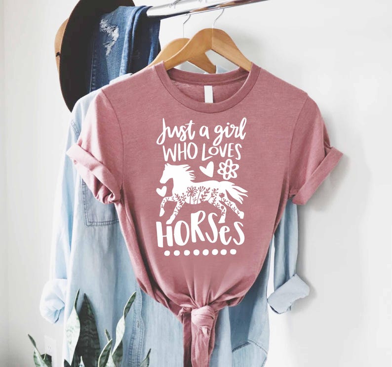 May include: Dusty rose t-shirt with the text "Just a girl who loves horses" and a white graphic of a horse. The horse design includes floral details. The shirt is hanging on a wooden hanger.