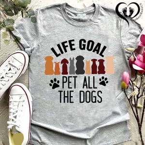 May include: A gray t-shirt with the text "LIFE GOAL PET ALL THE DOGS" in black. Above the text are colorful dog silhouettes. White sneakers are visible on the left side of the image. The shirt is a crew neck.