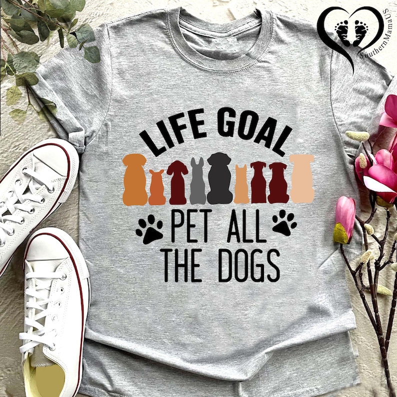 May include: A gray t-shirt with the text "LIFE GOAL PET ALL THE DOGS" in black. Above the text are colorful dog silhouettes. White sneakers are visible on the left side of the image. The shirt is a crew neck.