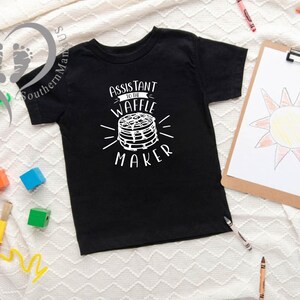 Puede incluir: Camiseta negra para ni&ntilde;os con el texto blanco "Assistant to the Waffle Maker". El gr&aacute;fico presenta una pila de gofres. Un dibujo de un sol est&aacute; en una tabla con pinza. Crayones y juguetes coloridos est&aacute;n dispersos.