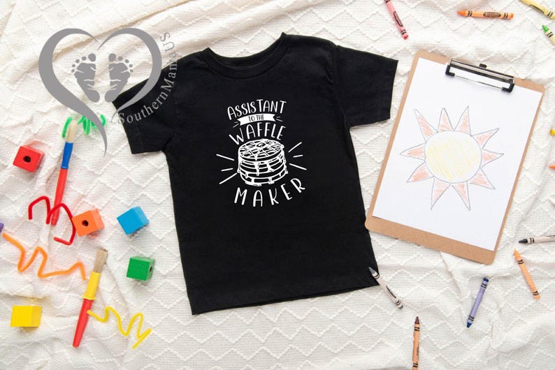 Puede incluir: Camiseta negra para ni&ntilde;os con el texto blanco "Assistant to the Waffle Maker". El gr&aacute;fico presenta una pila de gofres. Un dibujo de un sol est&aacute; en una tabla con pinza. Crayones y juguetes coloridos est&aacute;n dispersos.