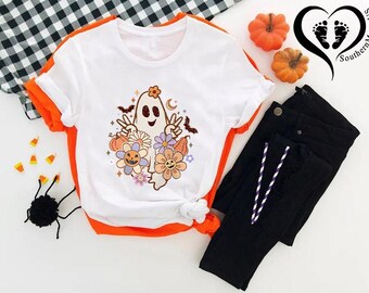May include: White t-shirt with an orange trim and a ghost graphic with flowers, bats, and a peace sign. Black pants with a drawstring, pumpkins, and a black and white checkered cloth. Halloween-themed apparel.