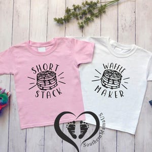 Puede incluir: Dos camisetas infantiles, una rosa y otra blanca, con dise&ntilde;os gr&aacute;ficos negros. La camiseta rosa dice "Short Stack" y la blanca "Waffle Maker". Ambas presentan una pila de panqueques.