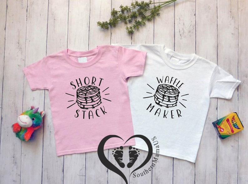 Puede incluir: Dos camisetas infantiles, una rosa y otra blanca, con dise&ntilde;os gr&aacute;ficos negros. La camiseta rosa dice "Short Stack" y la blanca "Waffle Maker". Ambas presentan una pila de panqueques.