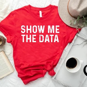 May include: A red t-shirt with the text "SHOW ME THE DATA" in white block letters. The shirt is displayed with a hat, a cup of coffee, a book, and a clock on a white surface.