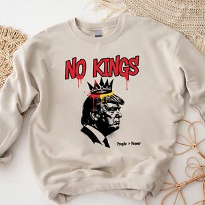 May include: A cream-colored sweatshirt with a graphic design. The design features a black and white profile of a person wearing a crown, with the words "NO KINGS" in red above. The phrase "People > Power" is printed below the profile.
