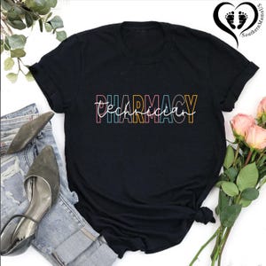K&ouml;nnte beinhalten: Schwarzes T-Shirt mit dem Schriftzug "Pharmacy Technician" in einer farbenfrohen, umrandeten Schriftart. Der Text ist eine Mischung aus Block- und Schreibschrift. Das Shirt wird mit Jeans, Abs&auml;tzen und Blumen gezeigt.