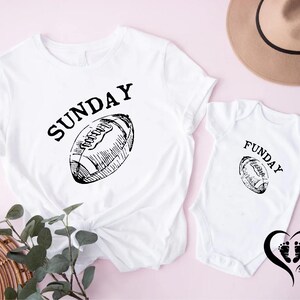 Puede incluir: Camiseta blanca con la palabra "SUNDAY" encima de un gr&aacute;fico de f&uacute;tbol americano. Un body de beb&eacute; blanco a juego con la palabra "FUNDAY" y un gr&aacute;fico de f&uacute;tbol americano. Un sombrero marr&oacute;n en el fondo.