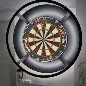 Könnte beinhalten: Eine Dartscheibe mit schwarzem Rahmen und einem schwarz-grauen Schrank. Die Dartscheibe hat ein traditionelles Design mit roten, grünen und beigen Segmenten. Drei Darts stecken in der Scheibe. Das Wort "Winmau" ist sichtbar.