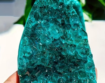 May include: A teal-colored crystal geode with a rough, textured surface. The crystal formation is visible, with many small, individual crystal structures. The geode is held in a hand, with a blurred green background.