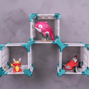 May include: A white and teal modular shelf unit with three connected cube shelves. Each shelf holds a different figurine: a pink fish, a red dragon, and a red race car. The shelves are mounted on a gray wall.