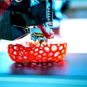 Könnte beinhalten: Ein 3D-Drucker erstellt ein rotes, filigranes, gitterartiges Objekt. Das Objekt wird auf einer blauen Oberfläche gedruckt.