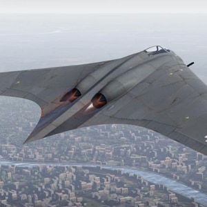 May include: A gray, wing-shaped aircraft with black and white markings flies over a city. The aircraft has two visible jet engines and a cockpit. The background shows a cityscape and a river.