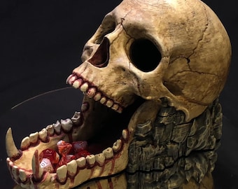 May include: A detailed, hand-painted skull dice tower with a gaping jaw and red-stained teeth. The skull is made of a beige resin and has a textured surface. The tower is designed to hold dice and features a compartment in the skull's mouth.