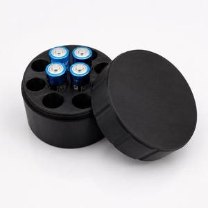 Könnte beinhalten: Ein runder, schwarzer Batterieaufbewahrungsbehälter aus Kunststoff mit abnehmbarem Deckel. Das Innere verfügt über acht kreisförmige Fächer, wobei vier blaue und silberne zylindrische Batterien in die vorgesehenen Plätze eingesetzt sind.