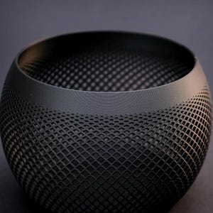 Op de afbeelding: Een zwarte, ronde kom met een textuur, roosterachtig patroon. De kom heeft een gladde rand en een donker interieur. Het ontwerp heeft een reeks kleine, gelijkmatig verdeelde openingen, waardoor een uniek visueel effect ontstaat. De kom is waarschijnlijk gemaakt van een stevig materiaal.
