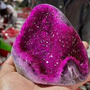 Könnte beinhalten: Eine polierte Amethyst-Geode mit tiefmagentafarbenem Innenraum und kristalliner Struktur. Der äußere Rand der Geode ist in einem helleren Violett gehalten, was einen auffälligen Kontrast erzeugt. Die Geode wird in einer Hand gehalten, um ihre Größe und Form zu zeigen.