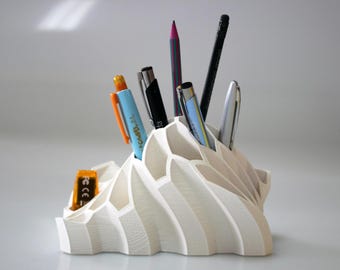 May include: White, 3D printed desk organizer shaped like a mountain range with multiple compartments. It holds a variety of pens, pencils, and a pencil sharpener.