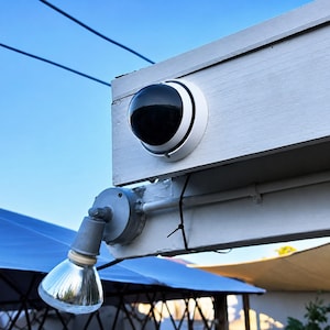 May include: A white and black security camera with a dome lens is mounted on a white wooden structure. Below, a silver spotlight is attached. The background shows a blue sky and a partially visible blue awning.