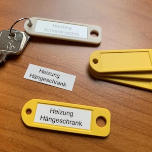 May include: A set of yellow key tags and a key on a wooden surface. The tags have white labels with German text. One tag is attached to a key ring. The text on the tags reads "Heizung Schrank rechts" and "Heizung Hängeschrank".