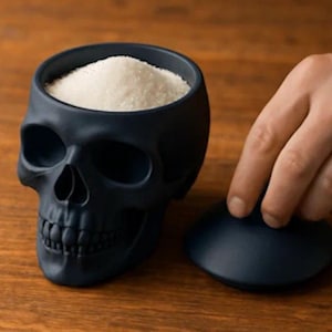 Könnte beinhalten: Ein schwarzer, schädelartiger Behälter, gefüllt mit weißen Granulaten, möglicherweise Salz oder Zucker. Der Behälter hat einen Deckel und steht auf einer Holzoberfläche. Eine Hand öffnet den Behälter.