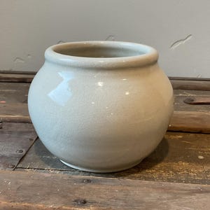 May include: A white ceramic pot with a rounded body and a slightly flared rim. The pot is sitting on a wooden surface.