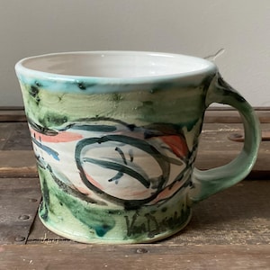Może przedstawiać: Ręcznie robiony ceramiczny kubek w zielono-niebiesko-białej kolorystyce. Kubek posiada abstrakcyjne malowane wzory w odcieniach zieleni, pomarańczy i czerni. Uchwyt i obręcz są w kolorze jasnozielonym. Kubek stoi na drewnianej powierzchni.