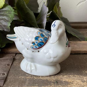 Może przedstawiać: Biała ceramiczna figurka kaczki z niebieskimi i złotymi detalami skrzydeł. Kaczka ma wzór w kształcie serca na głowie i małe otwory z boku. Figurka stoi na drewnianej powierzchni.