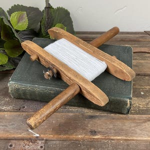 May include: A wooden hand-held kite string winder featuring a central spool wrapped with white string. The tool has two parallel wooden side bars connected by a central rod and a small metal wing nut fastener on one side of the wooden frame.