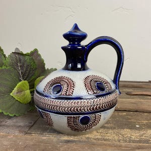 May include: A ceramic pitcher with a dark blue handle and a matching finial. The body is light gray with a repeating pattern of brown and dark blue swirls. The pitcher sits on a wooden surface.