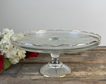 May include: A clear glass cake stand with a scalloped edge and gold trim. The stand has a round base and a pedestal. The cake stand is on a wooden surface.