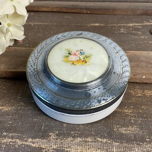 May include: A round, silver-toned trinket box with a white base. The lid features a painted scene of two figures in a garden. The box has a textured, patterned rim and a glossy finish. A vintage decorative item.