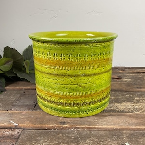 May include: A cylindrical ceramic planter featuring a vibrant lime green glaze with speckled texture. The exterior is decorated with horizontal bands of embossed geometric patterns and subtle orange-yellow accents around the center.