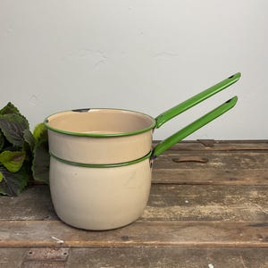 May include: Cream-colored enamel saucepan with a green rim and two green handles. The vintage pot has a rounded base and a smaller upper section. The pot is set against a rustic wooden surface.