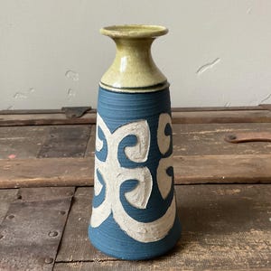 May include: A ceramic vase with a blue body and a cream-colored top. The blue section features a white, raised swirl design. The vase has a flared rim and a cylindrical shape, suitable for displaying flowers or as a decorative piece.