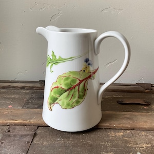 Op de afbeelding: Witte keramische kan met een gebogen handvat en tuit. De kan is versierd met illustraties van groene en rode bladeren. De kan staat op een houten oppervlak.