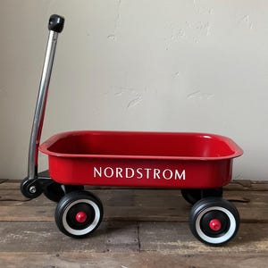 May include: A traditional red wagon with black wheels and a silver handle. The wagon features the word "NORDSTROM" in white. The wheels have white and red detailing.