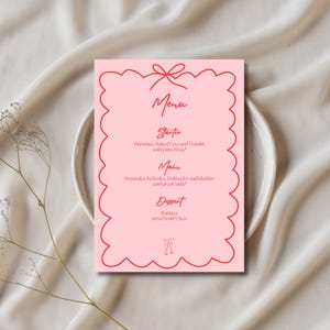 Peut inclure: Une carte de menu rose avec une bordure rouge festonnée et un motif de nœud. Le menu comprend des sections pour l'entrée, le plat principal et le dessert, avec du texte manuscrit en rouge. La carte est posée sur une assiette blanche.