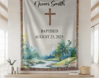 May include: A personalized baptism tapestry featuring the name "Noam Smith", a wooden cross, and the date "August 25, 2025". The tapestry has a watercolor landscape design with a river and trees, and a fringed border.