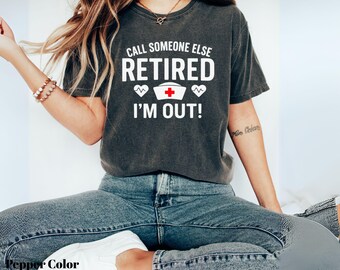 May include: A dark gray t-shirt with the text "CALL SOMEONE ELSE RETIRED I'M OUT!" in white. The design includes a nurse's hat with a red cross and two heart symbols. The shirt is worn with blue jeans.