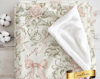 May include: A cream-colored blanket with a floral design featuring pink peonies, bows, and the name "Charlotte" in script. The blanket has a soft, white, plush interior and a gold ribbon with the word "Custom".
