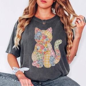 May include: Dark gray t-shirt featuring a patchwork cat design with floral, plaid, and solid fabric patches in various colors. The cat is centered on the shirt. The shirt has short sleeves and a crew neck.