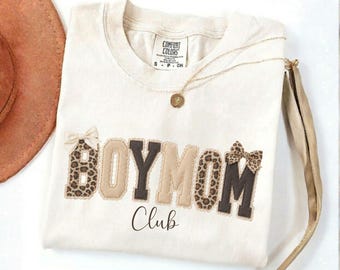 May include: A cream-colored t-shirt featuring the text BOY MOM in large, block-style letters with alternating leopard print and solid brown patterns. The word Club is printed in a cursive script below. Two small bows are attached to the letters.