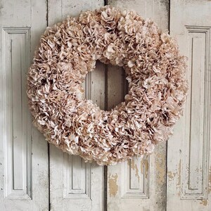 May include: A large, circular wreath made from many small, pale pink fabric flowers. It hangs against a distressed, white wooden door with vertical panels, giving a rustic appearance.