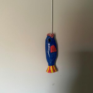 Peut inclure: Une tirette de lumière en forme de poisson en céramique bleue avec une finition brillante, dotée d'une nageoire rouge, d'un détail rose sur le dessus et d'une queue rayée jaune et rouge. Le poisson est fixé à un cordon blanc vertical.
