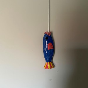 May include: A blue ceramic fish light pull with a glossy finish, featuring a red fin, a pink top detail, and a yellow and red striped tail. The fish is attached to a white cord that extends vertically from the top of the fish's head.